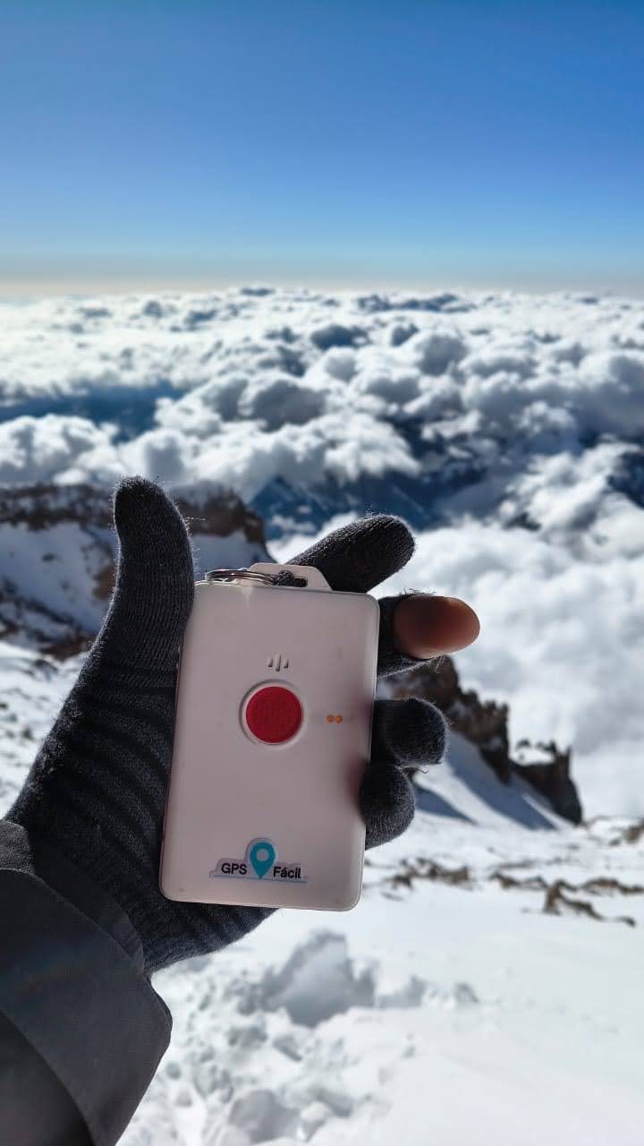 Una mano con guante sostiene un equipo GPS Fácil blanco con un botón rojo, en la cumbre del Aconcagua, sobre un mar de nubes y montañas nevadas.